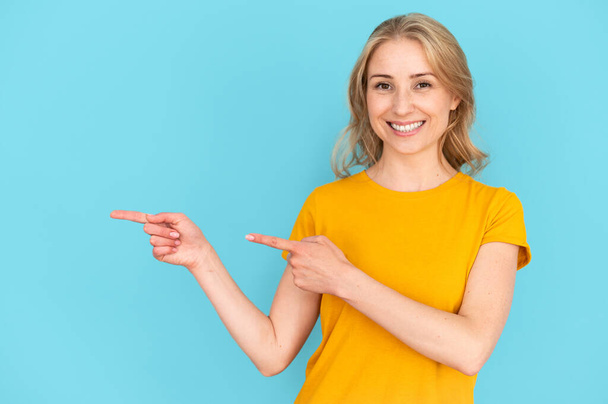 Female pointing with index fingers aside, showing empty place for advertising and shopping discount information. Happy woman in casual yellow t-shirt isolated on blue copy space background - Photo, Image