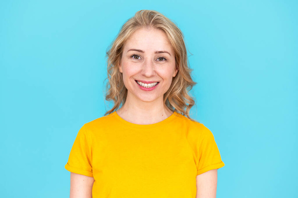 Portrét mladé ženy s příjemným úsměvem na modrém pozadí studia. Nadšená veselá žena hledící do kamery. Koncepce pózování a zobrazování bílých zdravých zubů, zubní ošetření - Fotografie, Obrázek