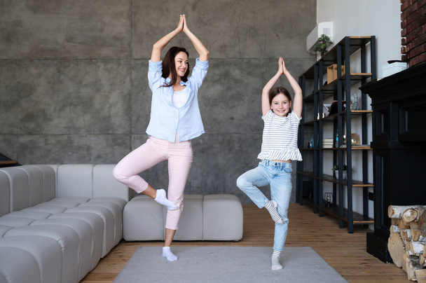 Gelukkige moeder die dochter leert om thuis yoga te doen. Vrouw en kind in boomhouding, glimlachend, genietend van vrijetijdsbesteding. Gezond levensstijl concept - Foto, afbeelding