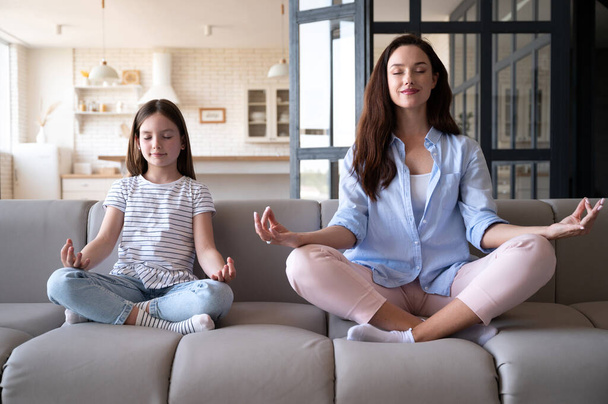 Calm young woman with preteen daughter sitting in lotus pose on couch and practice yoga together at home. Girl and mother meditation for relax and relieve negative emotions. - Photo, Image