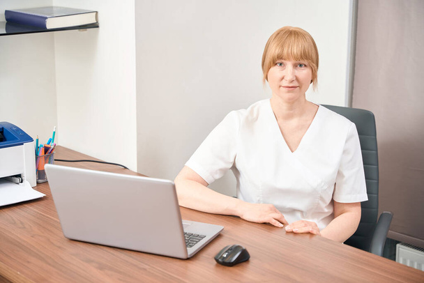 Hooggekwalificeerde vrouwelijke arts die wacht op een klant die met laptop op de gezellige werkplek zit, medische controles uitvoert, werkt in een privékliniek, gezondheidszorg en geneeskunde - Foto, afbeelding