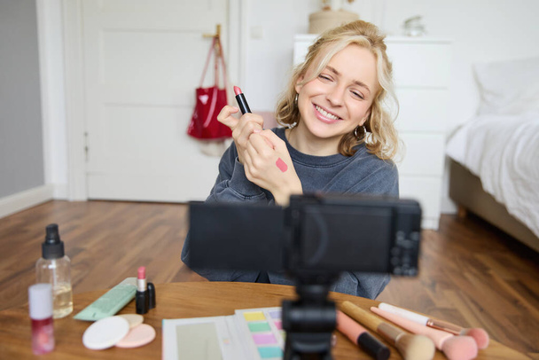 Portrait de jeune créateur de contenu de médias sociaux créatifs, femme montrant des échantillons de rouge à lèvres sur sa main, enregistrement vidéo sur la beauté et le maquillage, assis dans sa chambre devant un appareil photo numérique. - Photo, image