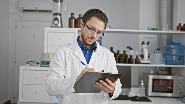 Young hispanic man scientist writing report standing at laboratory - Photo, Image