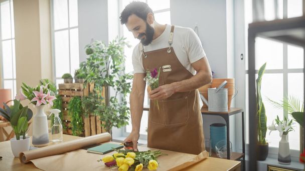 A bearded man in an apron arranges flowers in a sunny, plant-filled flower shop interior. - Foto, Bild
