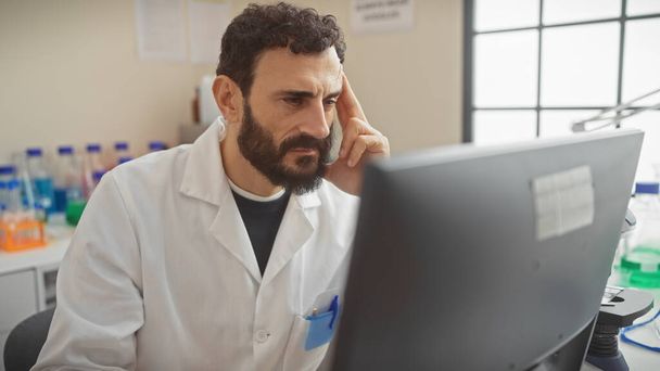 Hombre concentrado con barba usando bata de laboratorio analizando datos en computadora en laboratorio de investigación - Foto, Imagen