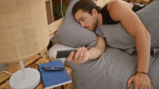 Hombre hispano joven usando teléfono inteligente acostado en la cama en el dormitorio - Foto, imagen