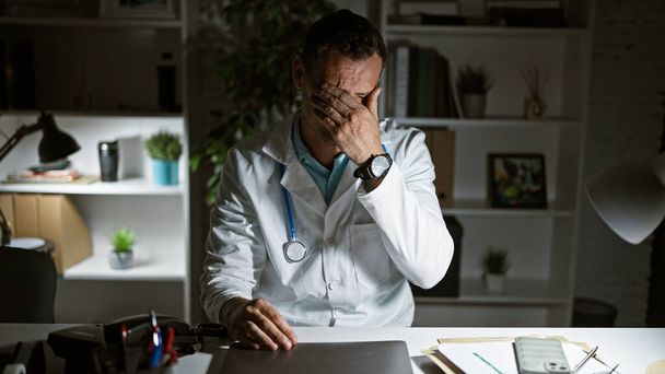Een gestresste man in een witte labjas zittend aan een bureau in een ziekenhuiskamer, die uitputting of bezorgdheid uitbeeldt. - Foto, afbeelding