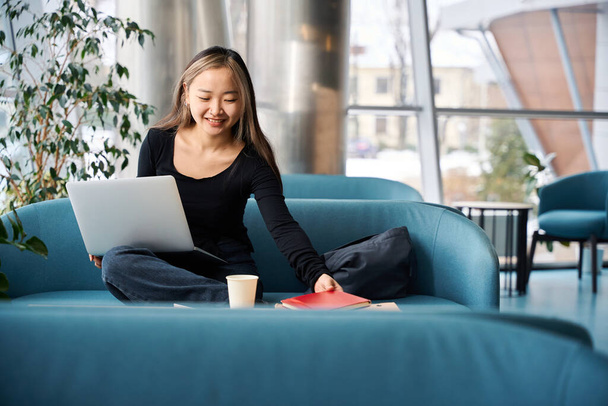 Nuori hymyilevä aasialainen IT-työntekijä ottaa kannettavan työskennellessään kannettavan tietokoneen sohvalla coworking toimistossa. Käsitteen moderni freelance tai etätyötä - Valokuva, kuva