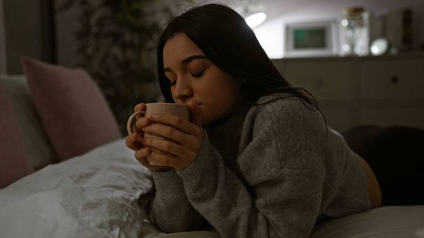 Hispanische Frau genießt einen ruhigen Moment mit einer Tasse drinnen, während einer gemütlichen, intimen Nacht zu Hause. - Foto, Bild