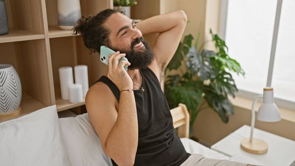 Iloinen latino mies puhuu puhelimessa viihtyisässä makuuhuoneessa, esittelee hänen rento tyyli ja moderni kodin sisustus. - Valokuva, kuva