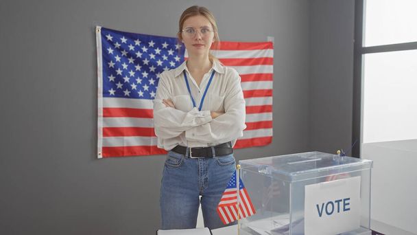 Mujer caucásica joven con los brazos cruzados se encuentra con confianza en una sala de colegio electoral con una urna y bandera. - Foto, imagen