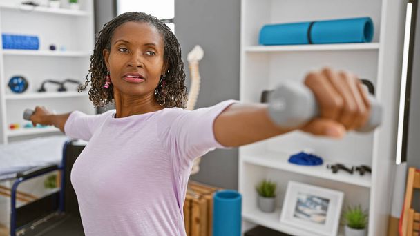 Mature african woman exercising with dumbbells in a rehab clinic interior - Photo, Image