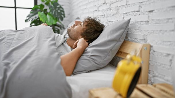 Ένας γενειοφόρος άνδρας αναπαύεται σε ένα μοντέρνο υπνοδωμάτιο με λευκούς τοίχους από τούβλα, δίπλα σε ένα ξυπνητήρι, προτείνοντας οπτικά ηρεμία και άνεση. - Φωτογραφία, εικόνα