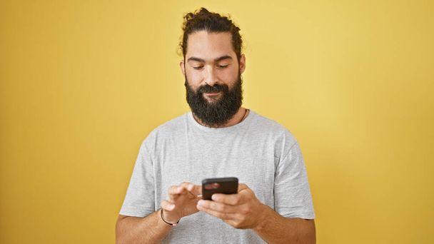 A young hispanic man with a beard smiles while using a smartphone against a yellow wall. - Φωτογραφία, εικόνα