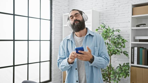 Modern bir ofiste rahat bir İspanyol adam akıllı telefon kullanırken kulaklıklarıyla müzik dinlemekten hoşlanır.. - Fotoğraf, Görsel