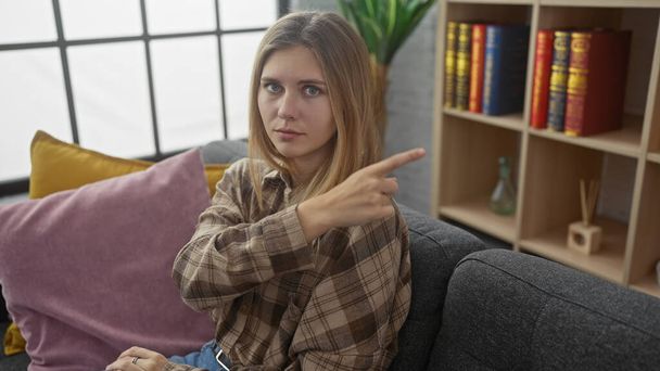 Młoda kobieta wskazuje siedząc na kanapie w nowoczesnym salonie, jej wyraz kontemplacyjny i poważny. - Zdjęcie, obraz
