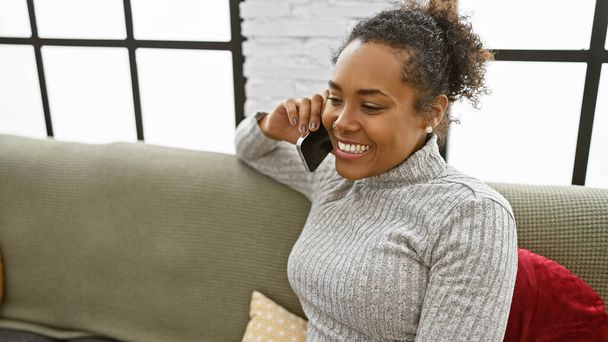 Una alegre mujer afroamericana hablando por teléfono en un acogedor salón. - Foto, Imagen
