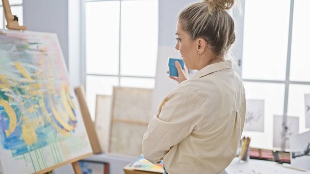 Молода блондинка художник дивиться малюнок тримає чашку кави в художній студії - Фото, зображення