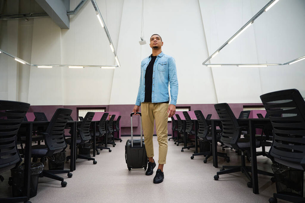 Полная фотография спокойного молодого менеджера, идущего по пустому офису с чемоданом - Фото, изображение
