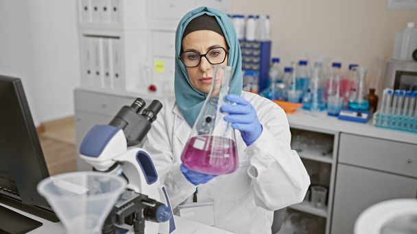 Μια στοχευμένη γυναίκα επιστήμονας εξετάζει ένα φλασκί σε ένα εργαστήριο, που περιβάλλεται από επιστημονικό εξοπλισμό και ένα μικροσκόπιο. - Φωτογραφία, εικόνα