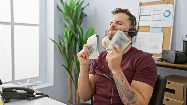 Un hombre hispano contento en una oficina se deleita con el dinero mientras usa auriculares. - Foto, imagen
