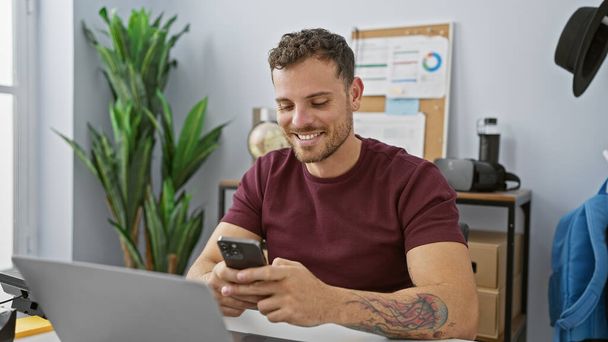 Uomo ispanico sorridente con barba utilizzando smartphone in ambiente ufficio moderno - Foto, immagini