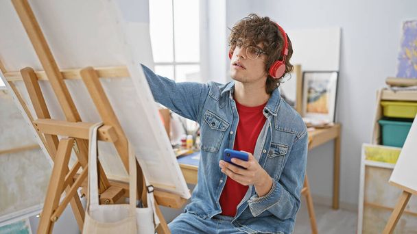 イーゼル絵とクリエイティブスタジオでスマートフォンを使用したヘッドフォンを持つ若い男. - 写真・画像