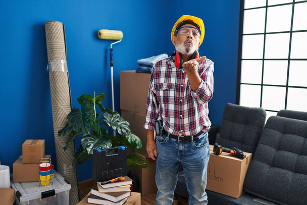 Cheeky senior man in hardhat and safety glasses blowing a love-filled kiss to the camera in his new home - a flirtatious handyman with a sparkle in his eye. - Photo, Image