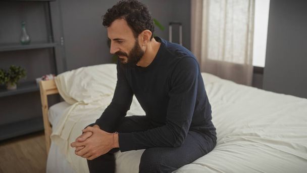 Ein nachdenklicher Mann mit Bart sitzt allein auf einem Bett in einem modernen Schlafzimmer und weckt Gefühle der Introspektion. - Foto, Bild