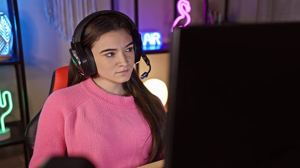 Focused young woman with headphones engages in gaming at home, showcasing a cozy, illuminated indoor environment. - Foto, imagen