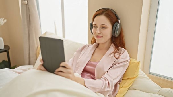 Een ontspannen vrouw met rood haar geniet van een ebook in een gezellige slaapkameromgeving, het dragen van een koptelefoon voor een meeslepende ervaring. - Foto, afbeelding