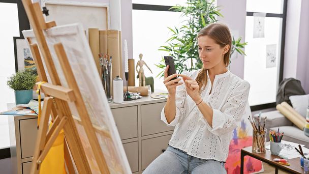 スタジオに焦点を当てた慎重な女性アーティストは,彼女のイーゼルとアート用品の横にスマートフォンを使用しています.. - 写真・画像