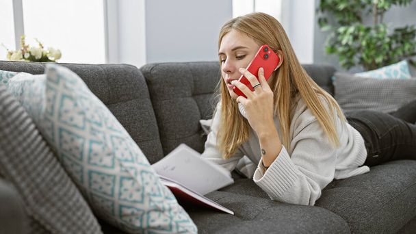 Blonde woman conversing on red smartphone lying comfortably on couch in modern living room setting - Фото, изображение
