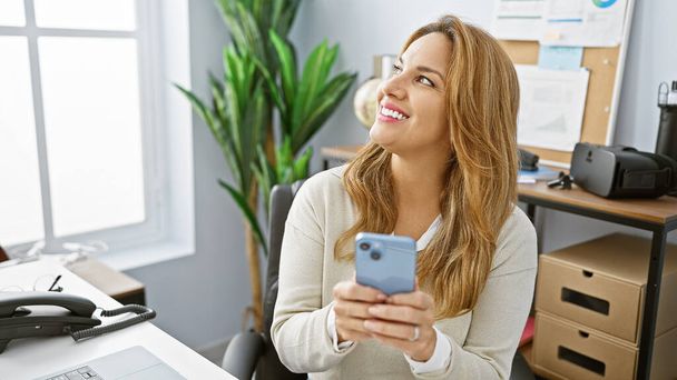 現代のオフィス環境でスマートフォンを使ったヒスパニック女性の笑顔. - 写真・画像