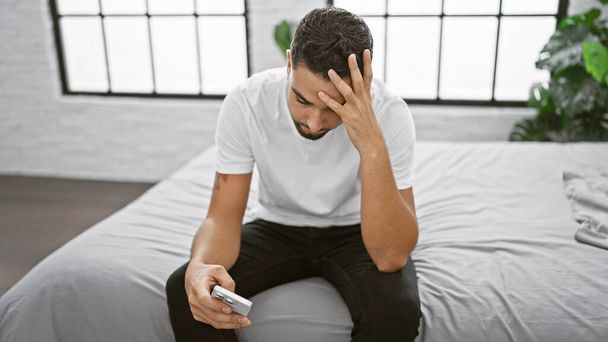 Gestresster junger Mann mit Bart und Smartphone im Schlafzimmer zeigt Ängste und Herausforderungen des modernen Lebensstils. - Foto, Bild