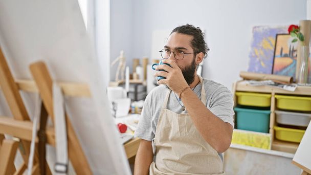 bel homme hispanique boire du café dans un studio d'art lumineux à l'intérieur - Photo, image