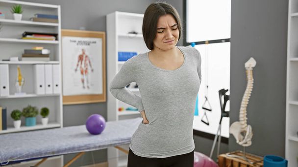 Eine junge hispanische Frau mit Rückenschmerzen steht in einer Physiotherapie-Klinik mit Anatomie-Postern und Geräten. - Foto, Bild
