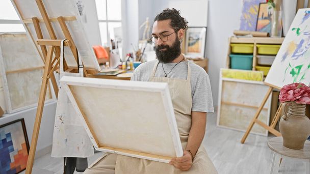 Un joven barbudo examina un lienzo vacío en un brillante estudio de arte lleno de caballetes y pinturas. - Foto, Imagen
