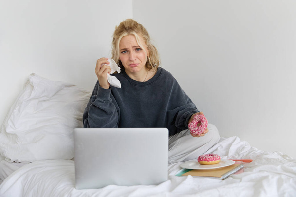 Portret van een vrouw die trieste film kijkt op laptop, huilt en tranen afveegt, donut eet en thee drinkt. - Foto, afbeelding