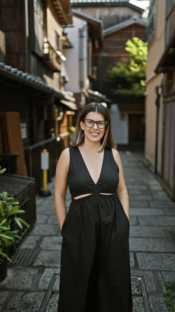 Mujer hispana segura, hermosa y alegre, posando alegremente con gafas puestas, de pie y sonriendo en las calles tradicionales de Kyoto, irradiando expresión latina natural y despreocupada. - Foto, imagen