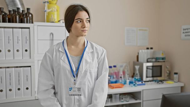 Una joven técnica hispana con bata de laboratorio se encuentra pensativamente en un ambiente de laboratorio, insinuando profesionalismo médico. - Foto, Imagen