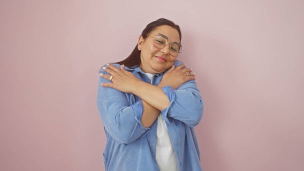 Латиноамериканка средних лет улыбается, обнимаясь с розовой стеной, вызывая тепло и позитив.. - Фото, изображение