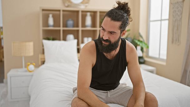 Un hombre hispano guapo con barba sonriendo en un dormitorio bien iluminado, posando casualmente en una cama. - Foto, imagen
