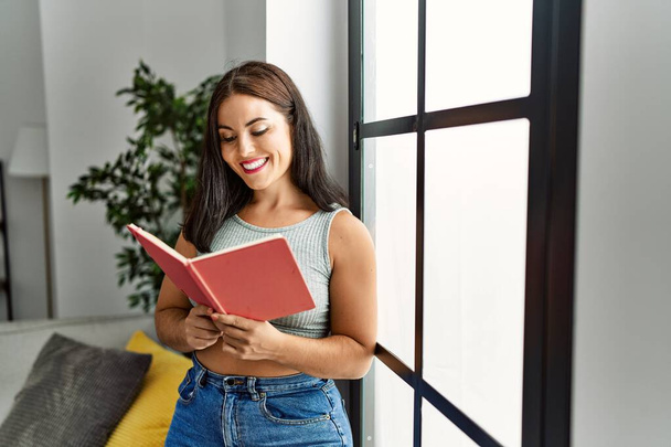 Молода красива іспаномовна жінка посміхається впевнено читаючи книгу вдома
 - Фото, зображення