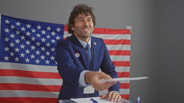 Mladý hispánec s plnovousem, oblečený v obleku, rozšiřuje volební lístek na americkou volební univerzitu zdobenou vlajkou.. - Fotografie, Obrázek