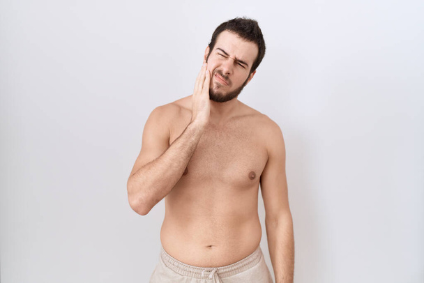 Jonge Spaanse man staat shirtloos over witte achtergrond raken mond met de hand met pijnlijke uitdrukking als gevolg van kiespijn of tandheelkundige ziekte op tanden. tandarts  - Foto, afbeelding