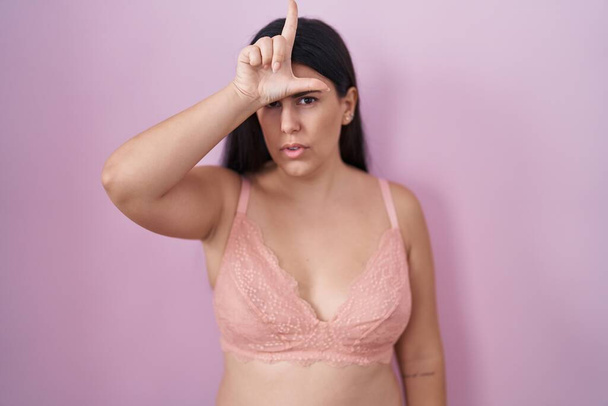 Young hispanic woman wearing pink bra making fun of people with fingers on forehead doing loser gesture mocking and insulting.  - Photo, Image