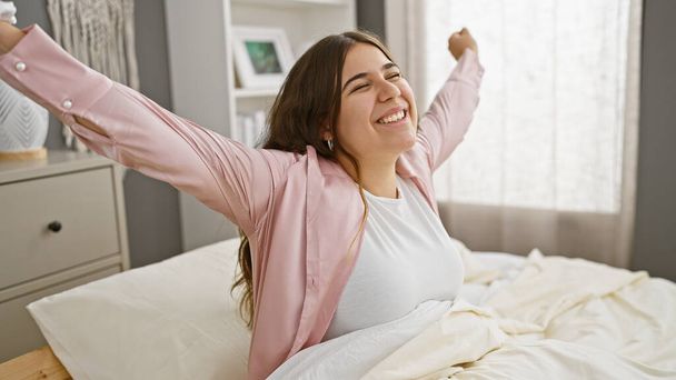 Une jeune femme joyeuse s'étire assise sur un lit, incarnant confort et bonheur dans un intérieur lumineux de chambre à coucher. - Photo, image