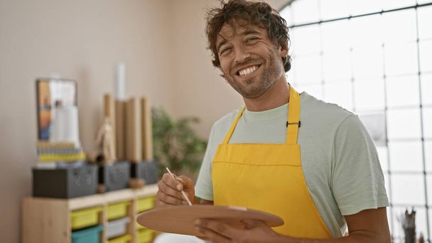 Homme barbu souriant dans le tablier tenant la palette dans le studio d'art ensoleillé - Photo, image
