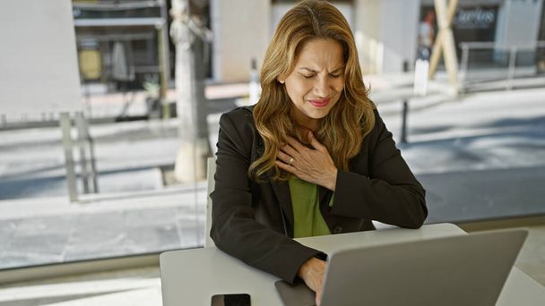 Hispanische Frau leidet an ihrem Arbeitsplatz im Büro unter Brustschmerzen, was auf mögliche gesundheitliche Probleme hinweist. - Foto, Bild